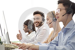 Happy male call centre operator showing thumb up gesture, working with his colleages at modern office