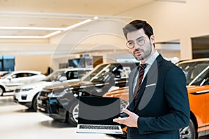 happy male car dealer in eyeglasses pointing at laptop with blank screen
