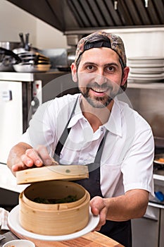 A happy male chef in a restaurant kitchen holding a bamboo dim sum steamer and smiling.