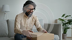 Happy male customer client consumer Arabian indian man guy in glasses open cardboard box at home unpack online shopping