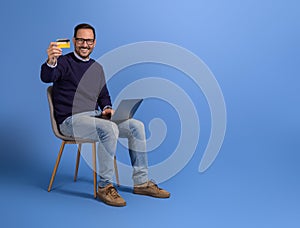 Happy male customer showing credit card and shopping over laptop on chair against blue background