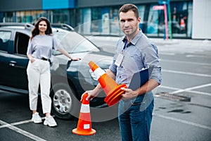 Happy male driving instructor holding traffic cones in his hands, looking at camera and smiling. Young woman standing