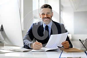 Happy male entrepreneur checking financial reports at desk in office and smiling at camera, enjoying company growth