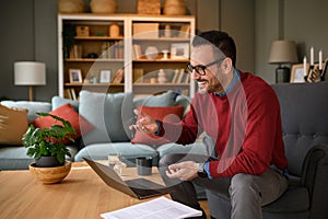 Happy male entrepreneur sitting on armchair and sharing ideas over online meeting on laptop at home