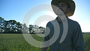 Happy male farmer in hat walking over green wheat field and enjoying nature. Young man going on cereal plantation and