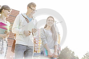 Happy male and female friends with books talking in university against sky