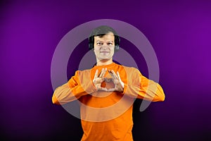 Happy male in headphones shows the gesture of the heart. A smiling guy in an orange T-shirt on a purple background shows a heart