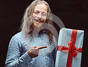 Happy male with long hair and beard in blue shirt pointing on gift box in his hand. Christmas concept.