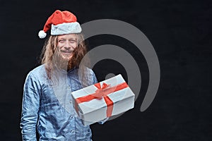 Happy male with long hair and beard in blue shirt and red Santa hat holds gift box.