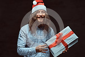 Happy male with long hair and beard in blue shirt and red Santa hat holds gift box.