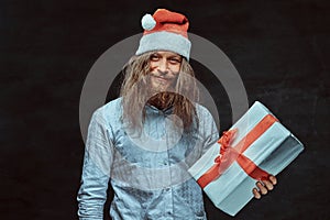 Happy male with long hair and beard in blue shirt and red Santa hat holds gift box.