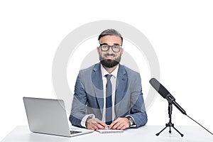 happy male newscaster sitting at table with laptop, notepad and microphone, photo