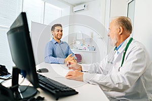 Happy male patient talking with mature adult doctor sitting at table during checkup visit in medical clinic office.