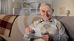 Happy male pensioner looking at photo sitting home sofa, good memories, feeling