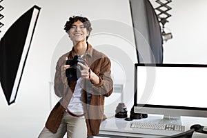 Happy male photographer with professional camera sitting on his desk with computer monitor with blank screen, mockup