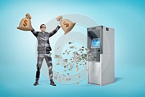 Happy male robber holding money bags with dollars falling out of ATM machine on blue background