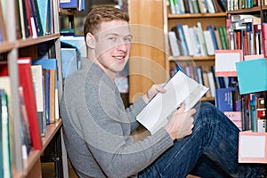 Happy male student with book sitting on floor in library