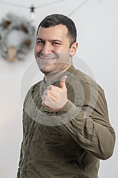 A happy man 30-35 years old holds a thumbs up against the background of a Christmas wreath. Concept: everything is cool, super hol