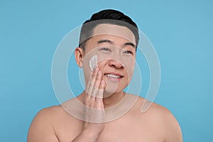 Happy man applying cream onto his face on light blue background