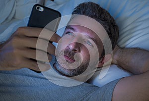 Happy man with blue eyes lying on bed late at night in dark light networking on mobile phone or online dating smiling relaxed