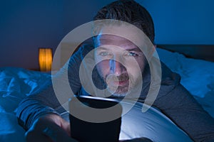 Happy man with blue eyes lying on bed late at night in dark light networking on mobile phone or online dating smiling relaxed