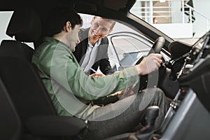 Happy man buying new vehicle, clarifying car purchase details with salesman, sitting inside auto salon at dealership