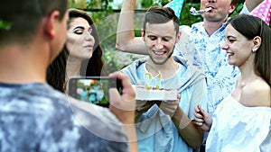 Happy man celebrates a birthday, blows out candles on a cake, friends congratulate, take a photo on mobile phone