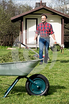 Happy man in a checked shirt carries a full of grass lawnmower container to emptying it into a wheelbarrow