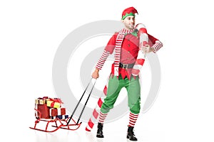 happy man in christmas elf costume carrying sleigh with presents and big candy cane isolated