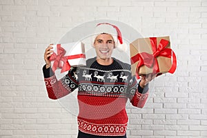 Happy man in  sweater and Santa hat holding gift boxes near white brick wall