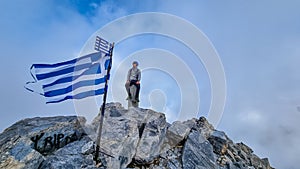 Happy man with climbing helmet on cloud covered mountain summit of Mytikas Mount Olympus, Greece. Greek flag on top