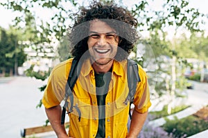 Happy man with curly hair smiling broadly, posing for social advertisement in the city street. Cheerful student male has joyful