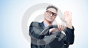 A happy man in a dark suit and glasses drives a car and sends greetings with his hand, on a light blue background.