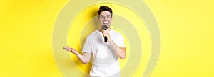 Happy man entertainer performing, talking in microphone, making speech or stand-up show, standing over yellow background