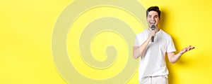 Happy man entertainer performing, talking in microphone, making speech or stand-up show, standing over yellow background