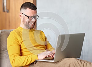 Happy man freelancer dressed casually using laptop while sitting in relaxed pose on sofa at home