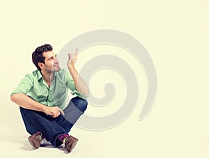 Happy man in green shirt and blue jeans sitting on the floor pointing at blank copy space