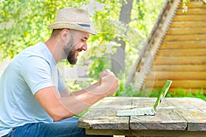 happy man in a hat with a beard, in a hat and a T-shirt sits at a wooden table in front of a laptop on a background of nature