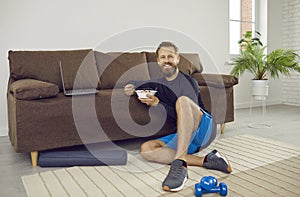 Happy man having break during his home workout, eating healthy food and using laptop