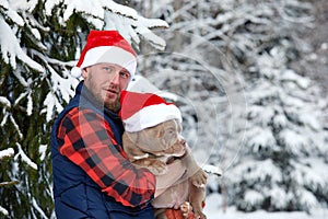 Happy man holding lovely dog in his hands wearing in a Santa hat in snowy forest. Smiling boy hugging adorable puppy in