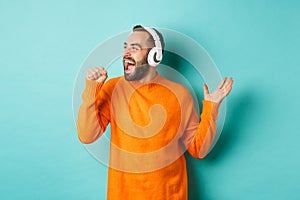 Happy man lipsync while listening music in headphones, holding invisible microphone, standing over turquoise background photo