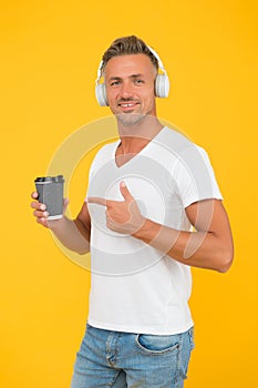 Happy man listen to music in modern headphones pointing finger at takeaway coffee cup yellow background, order