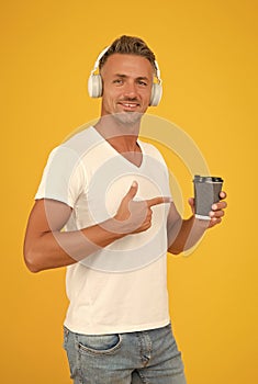 Happy man listen to music in modern headphones pointing finger at takeaway coffee cup yellow background, order