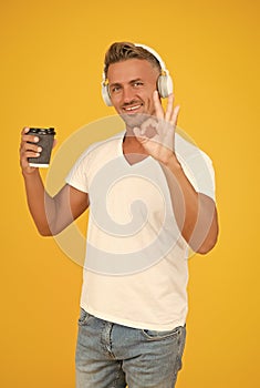 Happy man listen to music showing OK hand gesture for takeaway coffee in disposable cup yellow background, coffee-to-go