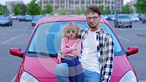 Happy man and little female kid in eyeglasses posing near red car on the parking while showing thumb up together. Family
