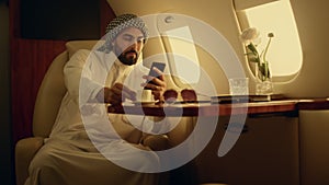 Happy man looking cellphone in private jet. Smiling arabian rest drinking coffee