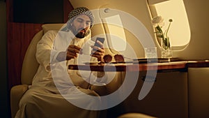 Happy man looking cellphone in private jet. Smiling arabian rest drinking coffee
