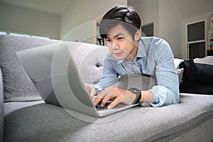 Happy man lying on sofa in living room and surfing internet with computer laptop.