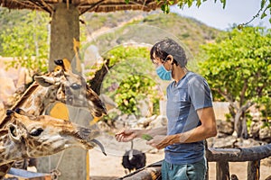 Happy man in medical mask watching and feeding giraffe in zoo. He having fun with animals safari park on warm summer day