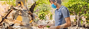 Happy man in medical mask watching and feeding giraffe in zoo. He having fun with animals safari park on warm summer day